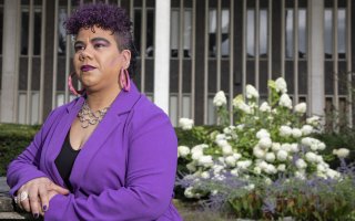 A woman in a purple jacket, with purple tinged hair and purple lipstick, stands in front of a hydrangea bush