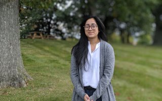 Thoin Begum, a PhD student, stands outside in a field, looking at the camera. Picnic tables and trees are in the background.