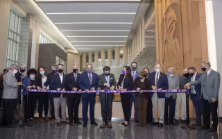 18 people in business clothes stand with scissors, ready to cut a purple ribbon stretched in front of them with the word ETEC repeated on it