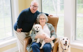 A smiling white-haired man leans over a silver-haired woman in a brown chair, with a white dog on her lap and another sitting beside her