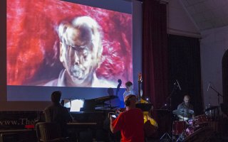 A large pink and blue photo-painting on a screen is on a stage before a group of piano-bass-drums musicians and a conductor