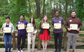 Six smiling students stand in a row in front of a wooden fence, each holding a scholarship certificate