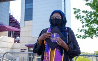 A soman wearing a headscarf, face mask and glasses along with graduation robes holds a circular sticker that says 1st generation graduate.