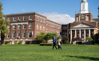 Two women walk across a lawn in front of two connected brick buildings, one with white pillars and a clock.