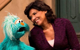 Sonia Manzano, in a purple sweater, leans in and smiles at a large, fuzzy, light blue Muppet