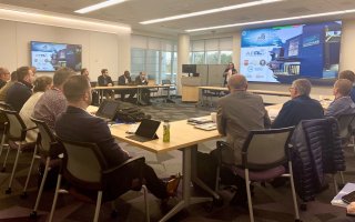 Attendees sit at tables at the circumference of a conference room as a woman standing near a projection screen speaks from the front of the room