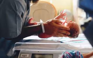 A person in gray medical scrubs and a hospital writband places a newborn infant on an electronic scale