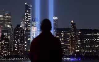 From the back, a silhoetted man stares at a nighttime city skyscape as two large blue beams of light reach to the sky