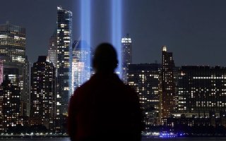 From the back, a silhoetted man stares at a nighttime city skyscape as two large blue beams of light reach to the sky