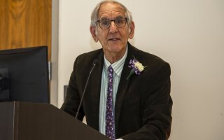 Professor Berman at a podium, wearing glasses, a brown jacket and purple tie, and a flower in his lapel.