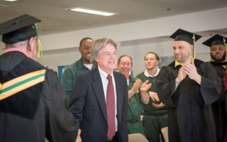 Randal Craig, smiling in a blue suit and red tie, is surrounded by clapping men in graduation garb or prison garb