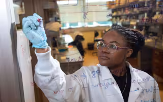 A woman in a lab coat and blue goves is seen behind a pane of glass she is writing formulas on.