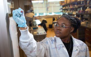 A woman in a lab coat and blue goves is seen behind a pane of glass she is writing formulas on.