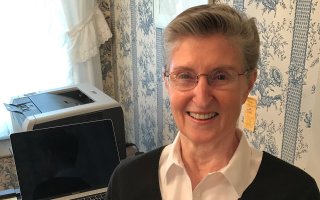 A woman smiles, sitting at a table holding her laptop computer and printer