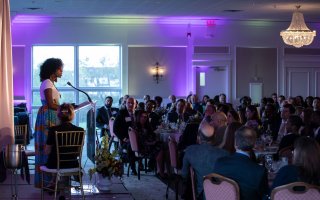 A woman in a long skirt speaks at a podium to a crowd seated at dinner tables