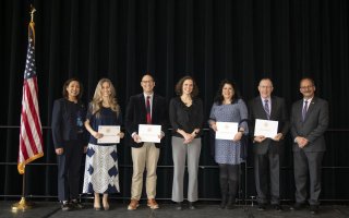 Seven smiling people, some holding awards certificate, stand in a row on a stage next to an American flag
