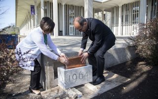 A man and a woman place a metal box into a cement cornerstone with 1964 engraved on it