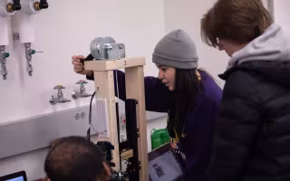 Engineering students at UAlbany work indoors on a rudimentary tensiometer device made of wood with a metallic gear at the top.