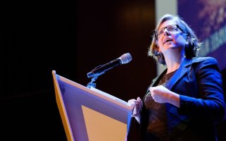 Virginia Eubanks, in glasses and a blue jacket, speaks at a microphone at a podium