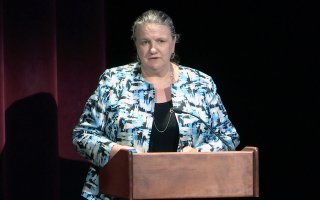 A woman stands at a podium with a red backdrop behind her wearing a blue and black jacket.