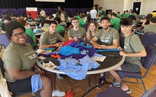 Six smiling UAlbany students in matching olive T-shirts are seated around a craft table as they pose for a picture.