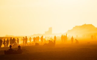 Beach crowded with people at sunset. The image is rich in orange and red tones; people are primarily cast as silhouettes. 