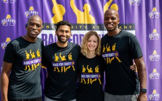 Four smiling adults wearing black "Race for Equity 5k" T-shirts stand in front of a purple fabric backdrop printed with University at Albany logos.