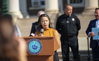 Associate Professor Tomoko Udo stands holding a microphone at a podium bearing the Albany County seal with several Albany County officials, including Sheriff Craig Apple, standing behind her as she speaks. 