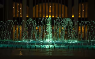 A fountain is lit with green lights, and seen in front of a building with glowing yellow lights from its windows