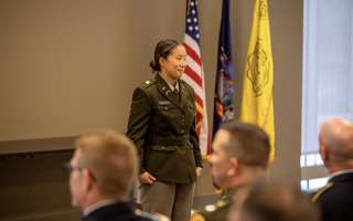A young woman in a U.S. Army uniform stands at attention in front of the U.S., Army and N.Y. State flags indoors with other officers in attendance.