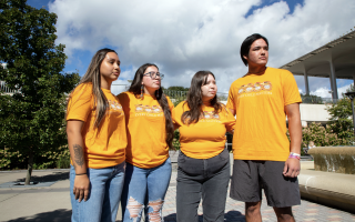 Four students in orange shirts that read "Every Child Matters" stare into the distance as they stand arm in arm next to a fountain on a sunny day. 