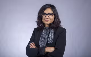 A portrait of Associate Professor Rukhsana Ahmed smiling with arms crossed in front of a plain gray background.