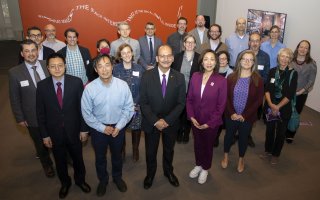 About two dozen faculty members stand together, hands clasped, looking up at the camera