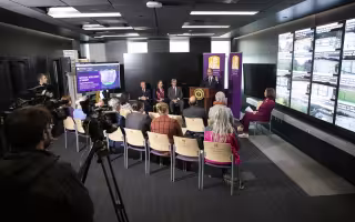 A wide-angle shot of a press conference in UAlbany's xCITE Lab. President Rodríguez stands at podium speaking flanked by two purple and gold UAlbany banners and 