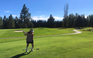 Kelly Gorman swings a gold club, teeing off, on a sunny day. The sky is bright blue with light cloud coverage and the grass is bright green and cut low. There are several sand traps in the background, as well as a cart path and trees around the edge of the golf course. She wears a white visor, white sneakers and pink sleeveless top. 