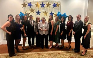 A group of 12 smiling people wearing formal attire stand in a row in front of a gauzy draped backdrop decorated with twinkle lights, gold and blue cutout stars, and gold and blue star shaped balloons.