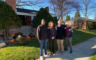 Four people in jackets stand on a sidewalk outside a brick house with a lawn decorated pumpkins.