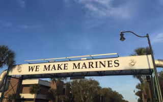 Sun illuminates a sign that reads "WE MAKE MARINES" against a backdrop of blue sky and palm trees in Parris Island, South Carolina.