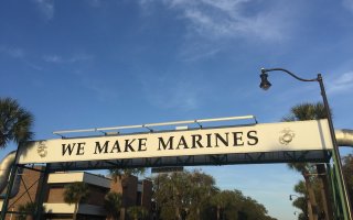 Sun illuminates a sign that reads "WE MAKE MARINES" against a backdrop of blue sky and palm trees in Parris Island, South Carolina.