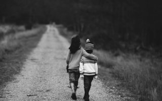 Two young children walk together down a wooded path; the taller child has their arm around the smaller one. 