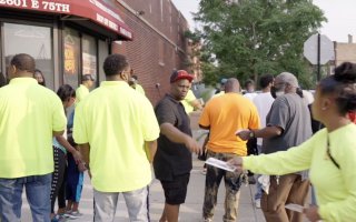 A group of people on a sidewalk. A woman in a yellow shirt hands a man a pamphlet as other men in yellow shirts stand nearby.