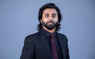 Portrait of a man with shoulder-length black hair and black beard, wearing a black suit jacket and purple tie, against a blue background.