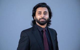 Portrait of a man with shoulder-length black hair and black beard, wearing a black suit jacket and purple tie, against a blue background.