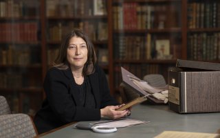 A woman with short brown hair dressed in black sits at a desk holding a gold leaf-trimmed book, a box of papers and a magnifying glass in front of her, and a glass case of books behind her.