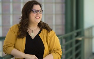 Dr. Melanie Sage wears a black top with yellow sweater, a gold necklace and purple rimmed glasses, leaning against a green railing indoors, smiling as she looks off to the side