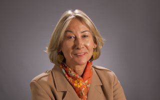 A woman with short blonde hair wears a camel jacket, patterned scarf and gold earrings and poses for a portrait.