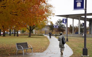 A person in a military uniform can be seen from behind walking along a path through Dutch Commons on the UAlbany campus on a wet fall day. A student walks toward them and trees display bright orange and yellow leaves.