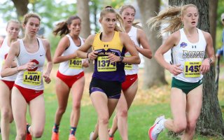 Cross country runners run through a wooded area with UAlbany XC Invitational bibs on their shirts.