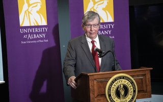 NYS Sen. Neil D. Breslin stands wearing a dark gray suit and red tie speaking at wooden lectern with the UAlbany seal framed on either side behind him by two purple UAlbany banners with the University's gold Minerva logo.