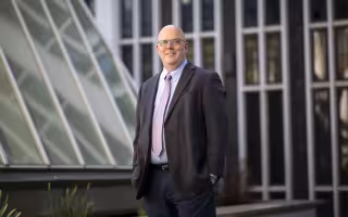 A man in a suit jacket and tie stands near glass windows